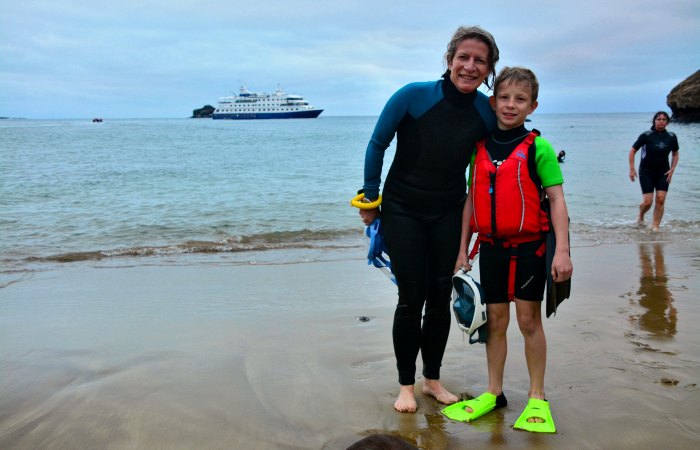 Galapagos family trip with ship in background and mother with son in wetsuits on the beach