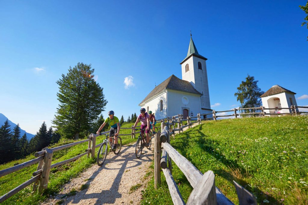 Slovenia family holidays - group cycling in Alpine scenery with traditional wooden church in background. Sky is blue.