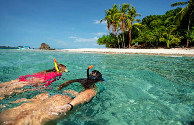 Family snorkelling in turquoise sea on Panama holiday with kids, with palm-fringed beach in the distance and blue sky
