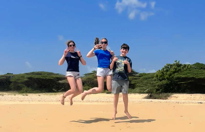 Teenagers with pre-teen sibling jumping together on Kenya's Indian Ocean coast - blue sky and golden sands